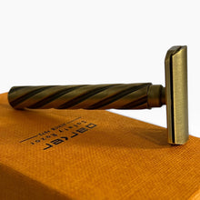Load image into Gallery viewer, A vintage-style safety razor with a twisted handle and brushed brass head, resting on an orange textured surface with a branded orange box behind it. The razor has a warm brass/gold finish with darker shadowing in the handle twist.
