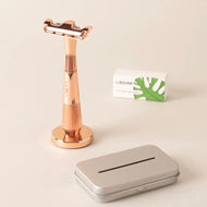 The image shows a rose gold safety razor kit. The kit includes a Leaf Twig Thorn Razor handle, a box of single-edge blades, a stand, and a recycling tin.